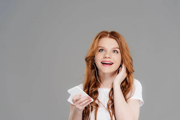 Beautiful smiling redhead girl in earphones holding smartphone and listening music isolated on grey with copy space — Stock Photo