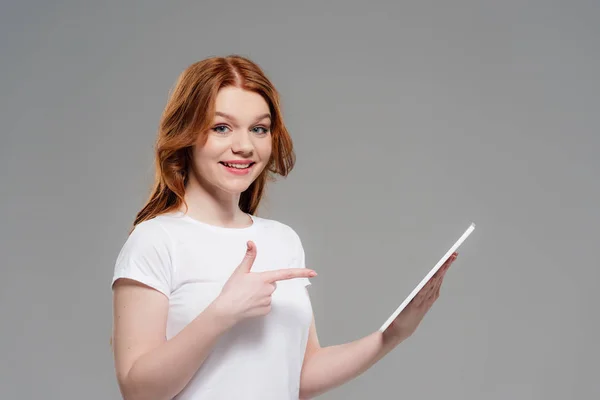 Schöne rothaarige Mädchen blickt in die Kamera, lächelt und zeigt mit dem Finger auf digitale Tablette isoliert auf grau — Stockfoto