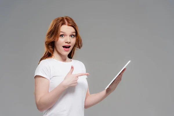 Beautiful surprised redhead girl looking at camera and pointing with finger at digital tablet isolated on grey — Stock Photo