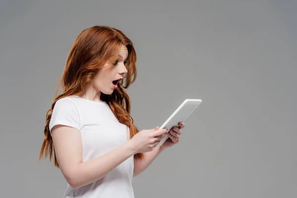 Красива здивована руда дівчина використовує цифровий планшет ізольована на сірому — стокове фото