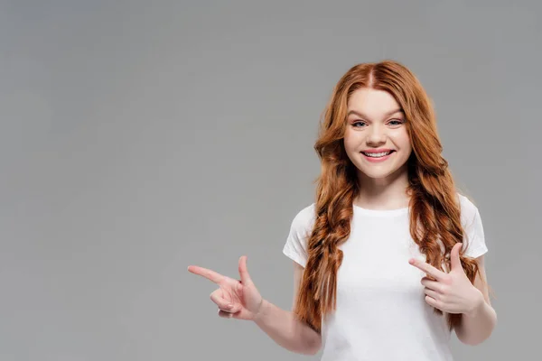 Beautiful redhead girl looking at camera, smiling and pointing with fingers isolated on grey with copy space — Stock Photo