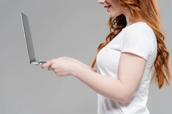 Обрізаний вид рудої дівчини в білій футболці з ноутбуком ізольовано на сірому — стокове фото