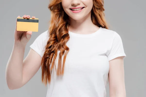 Обрезанный вид улыбающейся рыжей девушки, держащей кредитную карту с копировальным пространством, изолированным на сером — стоковое фото