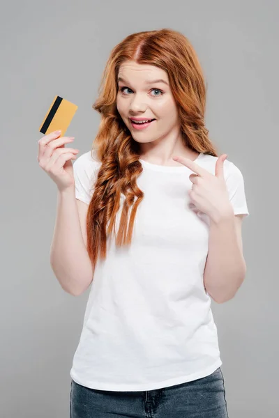 Belle rousse fille regardant caméra et pointant avec le doigt à la carte de crédit isolé sur gris — Photo de stock