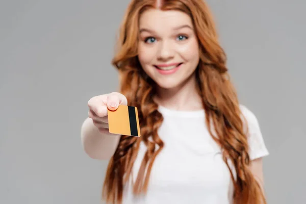 Hermosa chica pelirroja sonriente mirando a la cámara y mostrando la tarjeta de crédito aislado en gris - foto de stock