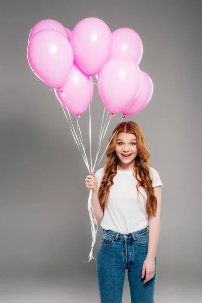 Hermosa chica sorprendida con el pelo rojo sosteniendo globos de aire rosa y mirando a la cámara en gris - foto de stock