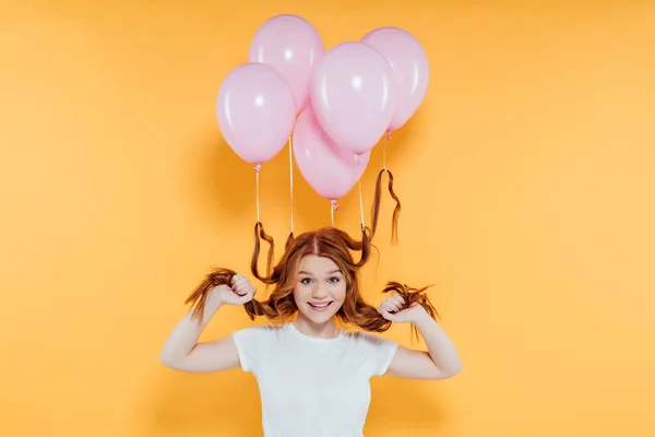 Happy redhead girl with balloons tied to hair posing isolated on yellow — Stock Photo