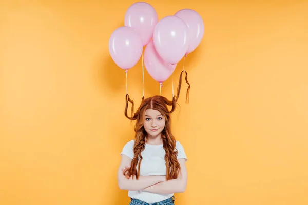 Rothaariges Mädchen mit zu Haaren gebundenen Luftballons blickt in die Kamera und posiert mit verschränkten Armen isoliert auf gelb — Stockfoto