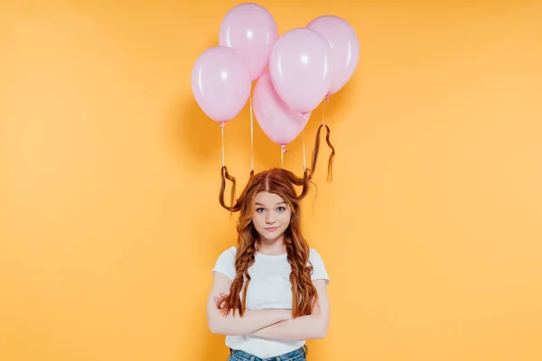 Pelirroja con globos atados al pelo mirando a la cámara y posando con los brazos cruzados aislados en amarillo - foto de stock