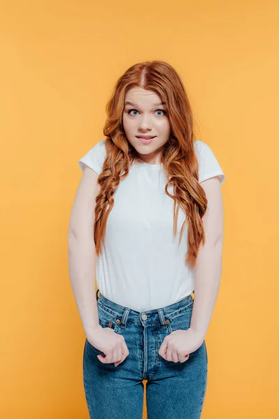 Beautiful redhead girl in white t-shirt and denim looking at camera and biting lip isolated on yellow — Stock Photo