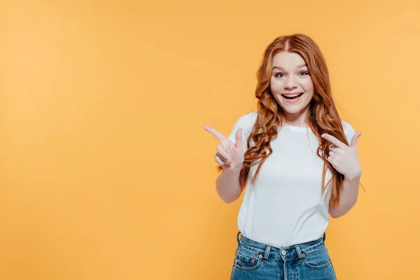 Schönes lächelndes rothaariges Mädchen, das in die Kamera schaut und mit den Fingern auf gelb mit Kopierraum zeigt — Stockfoto