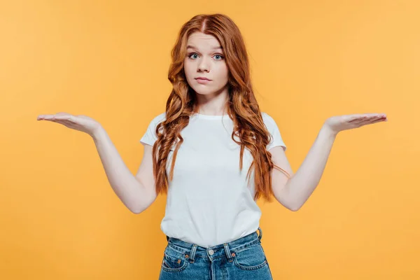 Beautiful redhead girl looking at camera and showing shrug gesture isolated on yellow — Stock Photo