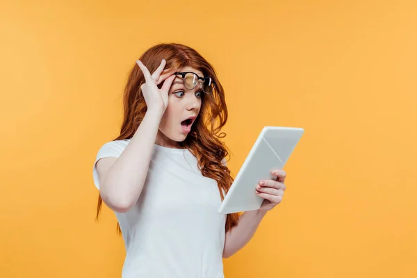 Surprised redhead girl with glasses using digital tablet isolated on yellow — Stock Photo