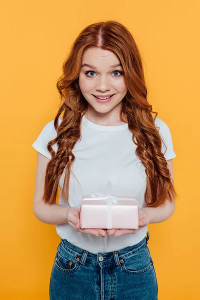 Beautiful redhead girl looking at camera and holding gift box isolated on yellow — Stock Photo