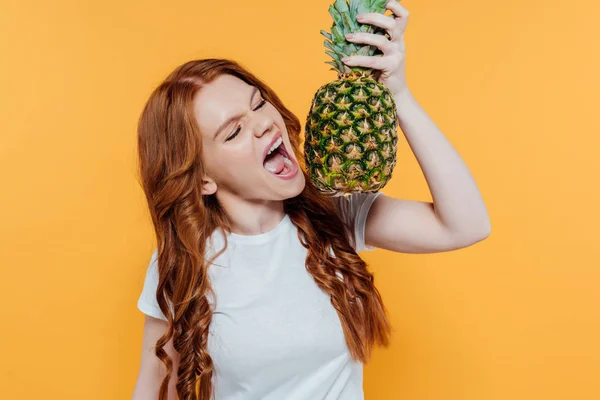 Beautiful redhead girl making face expression while biting pineapple isolated on yellow — Stock Photo