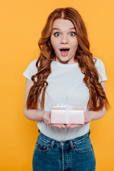 Überrascht rothaarige Mädchen schaut in die Kamera und hält Geschenkbox isoliert auf gelb — Stockfoto