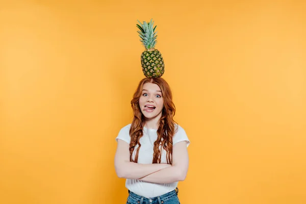 Redhead girl looking at camera, sticking tongue out and posing with pineapple on head isolated on yellow — Stock Photo