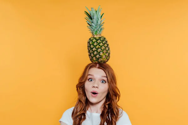 Surprised redhead girl looking at camera and posing with pineapple on head isolated on yellow — Stock Photo