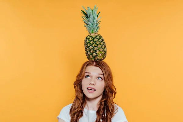 Rothaarige Mädchen schaut auf, während sie mit Ananas auf dem Kopf posiert, isoliert auf gelb — Stockfoto