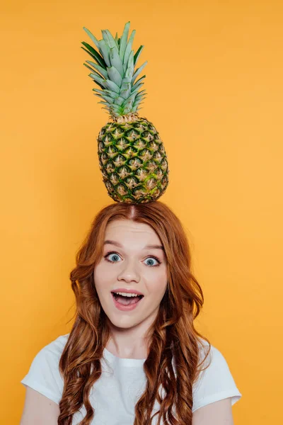 Excited redhead girl looking at camera and posing with pineapple on head isolated on yellow — Stock Photo