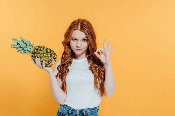 Bella rossa ragazza guardando la fotocamera e mostrando segno ok mentre posa con ananas isolato su giallo — Foto stock