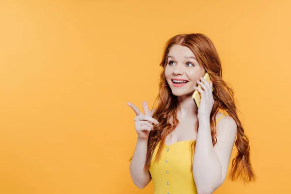 Belle fille rousse souriante pointant du doigt tout en parlant sur smartphone isolé sur jaune avec espace de copie — Photo de stock