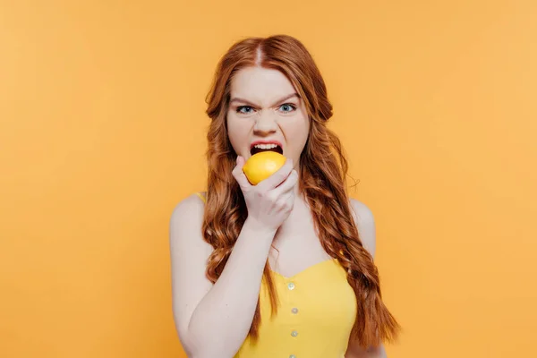 Redhead girl looking at camera, making facial expression and biting lemon isolated on yellow — Stock Photo