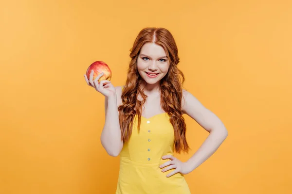 Красива усміхнена руда дівчина з яблуком дивиться на камеру ізольовано на жовтому — стокове фото