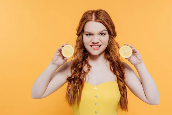 Pelirroja sosteniendo limones, mirando a la cámara y haciendo expresión facial aislada en amarillo - foto de stock