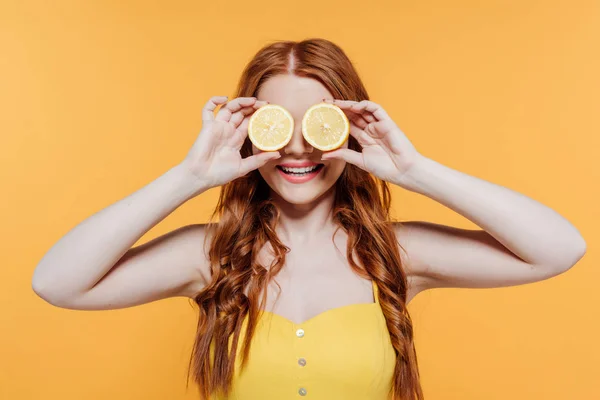 Redhead girl covering face with lemons and smiling isolated on yellow — Stock Photo