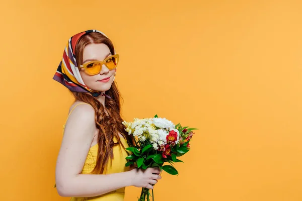 Bela menina elegante em lenço de cabeça e óculos de sol segurando flores isoladas no amarelo com espaço de cópia — Fotografia de Stock