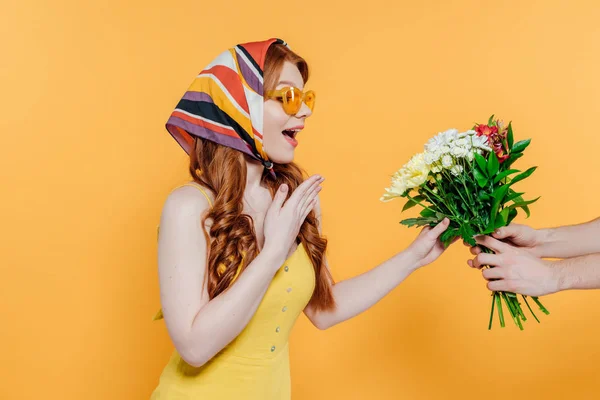 Überraschte stilvolle Mädchen mit Kopftuch und Sonnenbrille gestikulierend und mit Blick auf Blumen vereinzelt auf gelb — Stockfoto