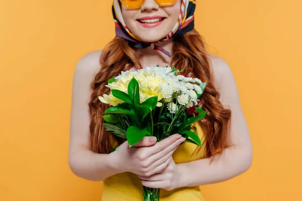 Обрезанный вид стильной рыжей девушки с цветами и улыбкой изолированы на желтый — стоковое фото