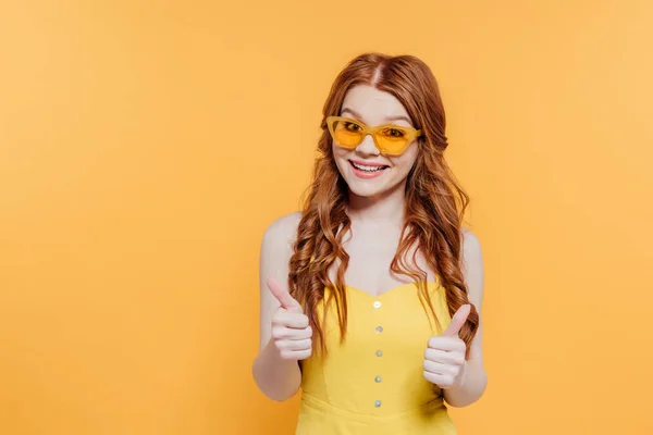 Belle rousse fille en lunettes de soleil regardant caméra et montrant pouces isolés sur jaune — Photo de stock