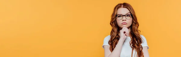 Panoramic shot of beautiful pensive redhead girl in glasses propping chin isolated on yellow with copy space — Stock Photo