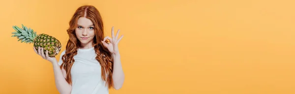Panoramic shot of beautiful redhead girl looking at camera and showing ok sign while posing with pineapple isolated on yellow — Stock Photo