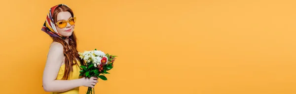 Tiro panorâmico de menina elegante bonita em lenço de cabeça e óculos de sol segurando flores isoladas no amarelo com espaço de cópia — Fotografia de Stock