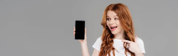 Colpo panoramico di bella ragazza rossa eccitato che punta con il dito allo smartphone con spazio copia isolato su grigio — Foto stock