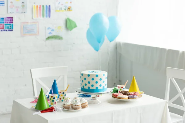 Bolo festivo e bonés de festa na mesa no quarto decorado para festa de aniversário com balões de ar — Fotografia de Stock