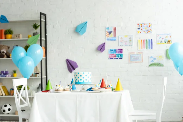 Festive cake and party caps on table in room decorated for birthday party with air balloons — Stock Photo