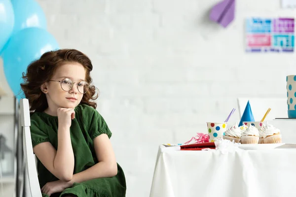 Sad kid sitting at table with cupcakes and propping chin during birthday celebration — Stock Photo