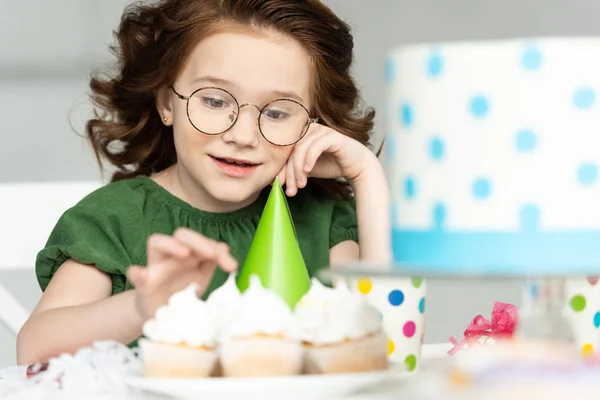Adorable preadolescente sentado en la mesa y mirando cupcakes durante la celebración del cumpleaños en casa - foto de stock