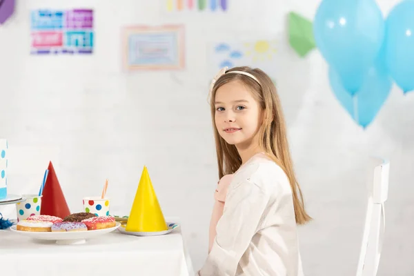 Очаровательный ребенок смотрит в камеру, сидя за столом с праздничными украшениями и пончиками во время празднования дня рождения — стоковое фото