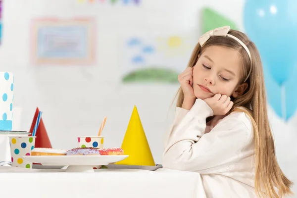 Enfant bouleversé assis à la table avec des beignets et des casquettes pendant la célébration de l'anniversaire — Photo de stock