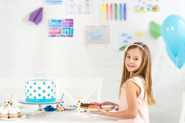 Adorable kid holding plate with donuts at birthday table with cake during party — Stock Photo