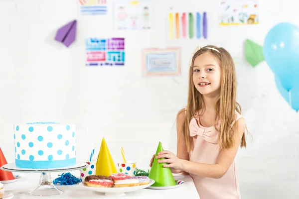 Очаровательный улыбающийся ребенок за праздничным столом с праздничными шапочками и праздничным тортом, смотрящим в камеру — стоковое фото