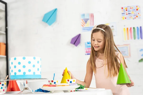 Adorable smiling kid at festive table with party caps and birthday cake — Stock Photo