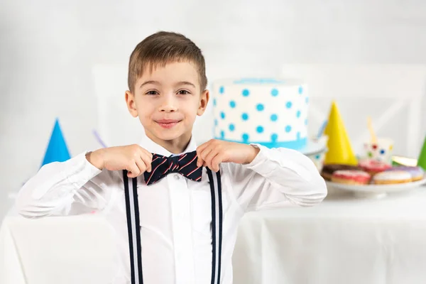 Adorable chico ajustando pajarita y mirando a cámara durante fiesta de cumpleaños - foto de stock