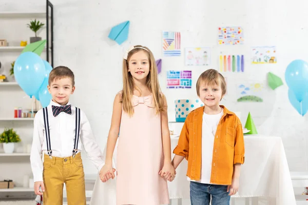 Adorables enfants regardant la caméra et tenant la main pendant la fête d'anniversaire à la maison — Photo de stock