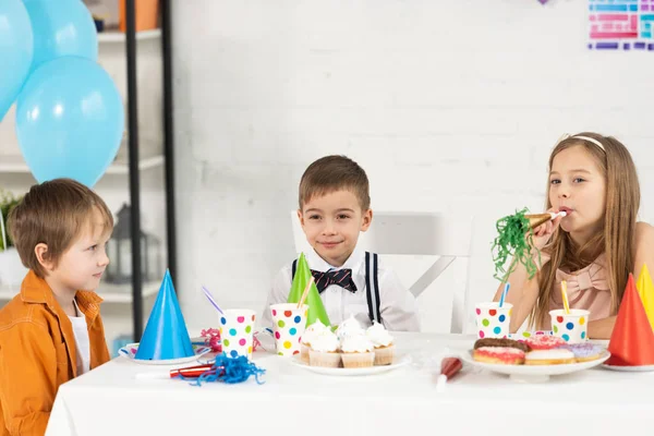 Crianças sentadas à mesa com cupcakes e chifres de festa durante a celebração do aniversário — Fotografia de Stock